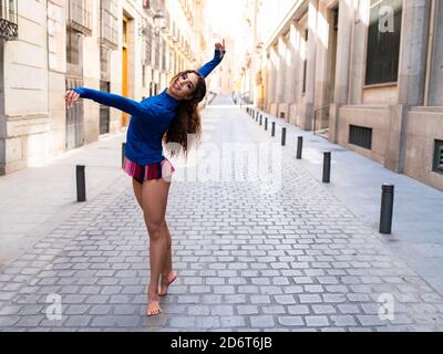 Vue latérale d'une jeune femme ethnique sportive et ébouriffée souriant tout en dansant dans la rue entre les anciens bâtiments en plein soleil Banque D'Images