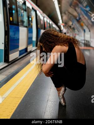 Vue latérale d'une jeune ballerine méconnue avec de longs cheveux bouclés dans les chaussures pointe embrassant les genoux tout en étant assis sur les haunches dedans station de métro Banque D'Images
