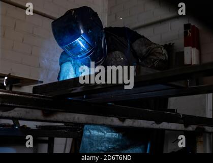 Employé masculin méconnaissable dans des gants de protection et un casque par soudage machine lors de travaux dans un atelier sombre Banque D'Images