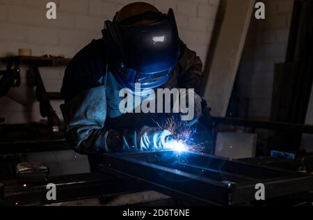 Employé masculin méconnaissable dans des gants de protection et un casque par soudage machine lors de travaux dans un atelier sombre Banque D'Images