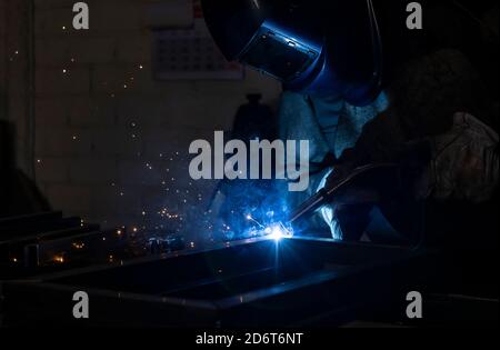 Employé masculin méconnaissable dans des gants de protection et un casque par soudage machine lors de travaux dans un atelier sombre Banque D'Images