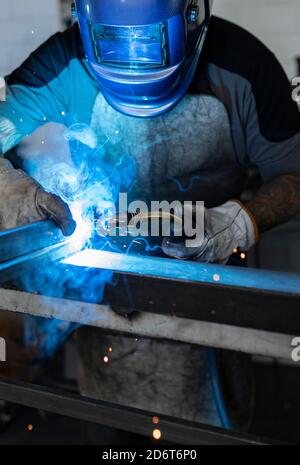 Employé masculin méconnaissable dans des gants de protection et un casque par soudage machine lors de travaux dans un atelier sombre Banque D'Images