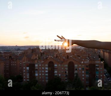 Crop femelle sans visage avec les bouts de pouce et d'index ensemble autour d'un soleil de soirée brillant au-dessus des bâtiments modernes de la ville Banque D'Images