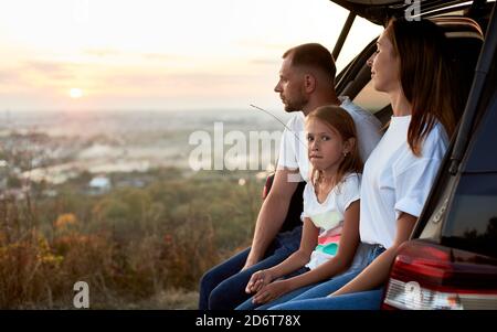 Vue latérale d'une jeune famille assise dans le coffre de voiture en profitant du coucher de soleil à l'extérieur de la ville, fille avec une paille dans sa bouche est regarder loin, espace de copie Banque D'Images