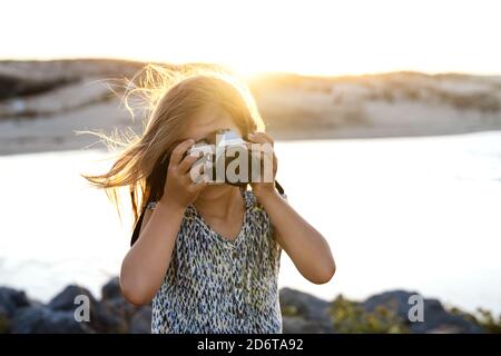 Jeune touriste femelle méconnaissable dans une robe élégante prenant photo de mer sur la rive rocheuse au coucher du soleil Banque D'Images