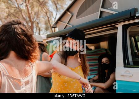 Femmes amicales dans des masques de protection debout près d'une camionnette dans la forêt et se cogner les coudes tout en se saluant les uns les autres pendant l'épidémie de coronavirus Banque D'Images