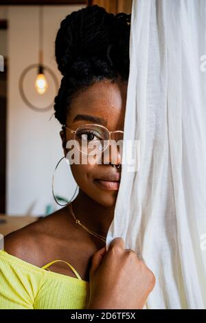 Jeune femme afro-américaine pensive en lunettes tendance couvrant la moitié du visage avec un rideau et regardant la caméra à la maison Banque D'Images