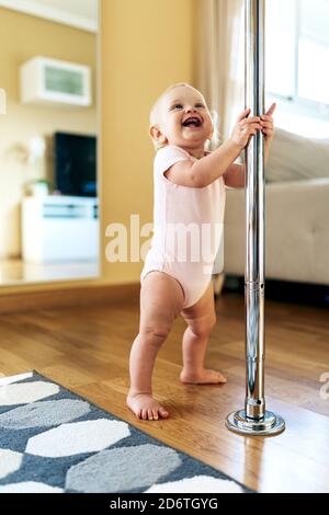 Pleine longueur de gaie barefooted petite fille dans bodysuit tenue perche métallique et rire tout en faisant les premiers pas à la maison Banque D'Images