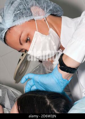 Dermatologue cosmetologiste visite patient avant la procédure de beauté ou le soulèvement de visage, belle femme médecin, robe avec capuchon chirurgical et masque avec chirurgie Banque D'Images