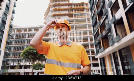 Employé barbu en uniforme orange mettant un casque sur sa tête tout en se tenant à l'extérieur du bâtiment pendant la construction. Construction mâle caucasienne Banque D'Images