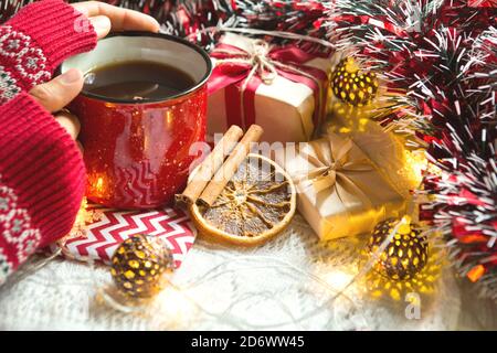 La main d'une femme dans un chandail chaud tient une tasse rouge avec une boisson chaude sur une table avec des décorations de Noël. Atmosphère du nouvel an, bâtons de cannelle et un Banque D'Images