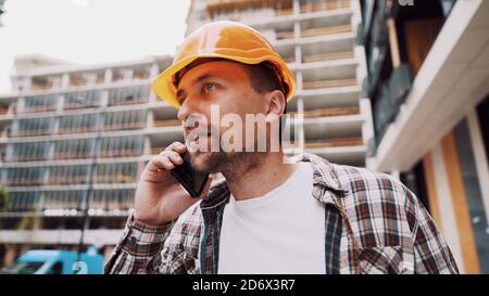 Un architecte mâle en colère dans un casque orange dur parle des oreilles au téléphone sur le chantier de construction. Le créateur en chemise à carreaux déplaisait émotionnellement négocier par cellule Banque D'Images