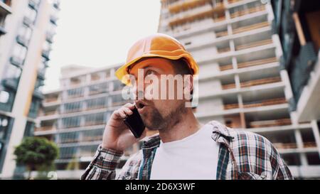 Un architecte mâle en colère dans un casque orange dur parle des oreilles au téléphone sur le chantier de construction. Le créateur en chemise à carreaux déplaisait émotionnellement négocier par cellule Banque D'Images