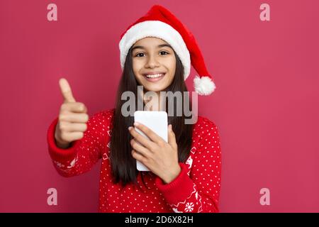 Bonne fille indienne porter chapeau de père noël tenir téléphone montrer les pouces sur fond rouge. Banque D'Images