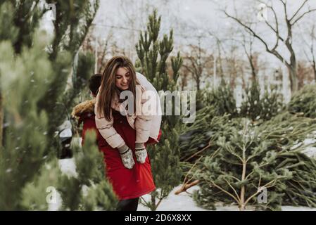 Un jeune couple se berce, s'amusant en hiver sur le fond du marché des arbres de Noël dans la ville. Une jeune femme sur le dos de son petit ami rit et sourit. Banque D'Images