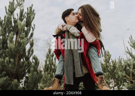 Un beau jeune couple est venu choisir un arbre. Les jeunes mariés amoureux se trompent en hiver. Une jolie femme rit sur les épaules d'un homme. Banque D'Images