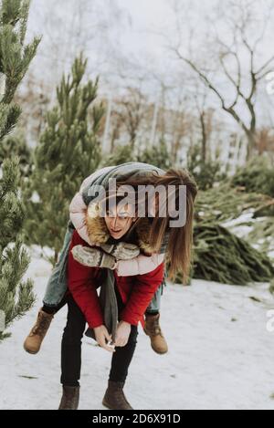 Un jeune couple se berce, s'amusant en hiver sur le fond du marché des arbres de Noël dans la ville. Une jeune femme sur le dos de son petit ami rit et sourit. Banque D'Images