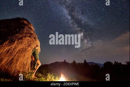 Une jeune femme près d'une botte de foin et d'un feu de camp sur fond de vallée de collines, sous un magnifique ciel étoilé de nuit sur lequel la voie lactée est visible. Banque D'Images