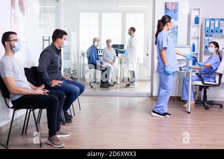 Une infirmière qui prend les radiographies d'un patient d'un autre personnel médical portant un masque facial contre le coronavirus dans la salle d'attente de l'hôpital. Assistant travaillant sur l'ordinateur de réception. Médecin dicusing avec couple senior Banque D'Images