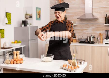 Homme âgé portant un tablier et bonete préparant une pizza savoureuse qui craque des œufs sur la farine. Chef pâtissier craquant l'œuf sur un bol en verre pour la recette du gâteau dans la cuisine, le mélange à la main, le pétrissage. Banque D'Images