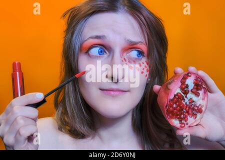 Jeune femme avec grenade et rouge à lèvres, gros plan. Rouge à lèvres et fruit rouge sur fond orange. Visage féminin et grenat rouge. Banque D'Images