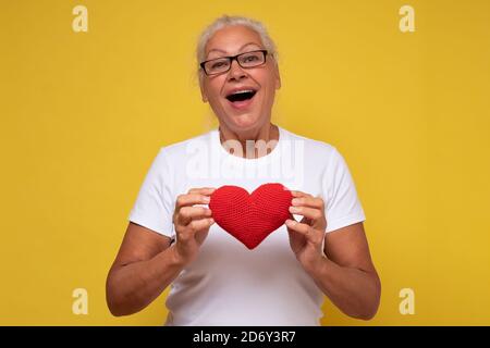 Femme hispanique senior avec des lunettes souriantes et amoureuses montrant le cœur rouge. Banque D'Images
