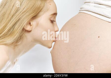 Une fille blanche blonde embrasse le ventre de sa mère enceinte isolée. Concept de famille et de sœur âgée, vue latérale. Banque D'Images