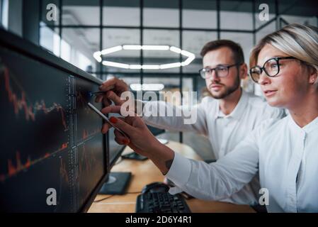 Place le stylet sur l'écran. Deux courtiers en vêtements officiels travaillent dans le bureau avec le marché financier Banque D'Images