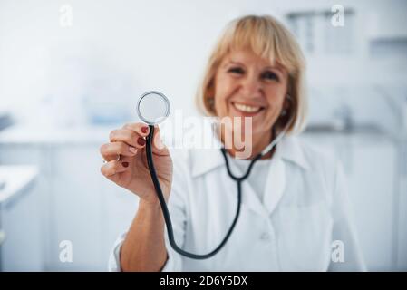 Tient le stéthoscope en main. Portrait d'un médecin senior en uniforme blanc qui se tient dans la clinique Banque D'Images