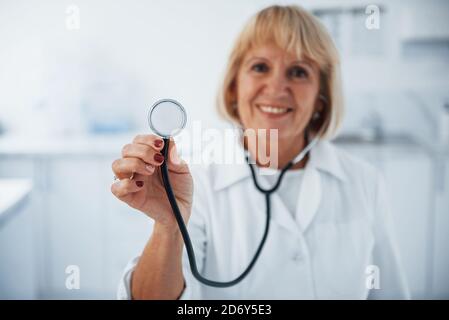 Tient le stéthoscope en main. Portrait d'un médecin senior en uniforme blanc qui se tient dans la clinique Banque D'Images