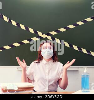 Une femme enseignante dans le masque médical jette ses mains dans la confusion. Le tableau noir est recouvert de ruban jaune et noir. Concept de problèmes avec educati Banque D'Images