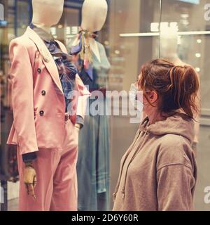 Une jeune femme dans un masque médical regarde le fenêtre d'un magasin de vêtements Banque D'Images