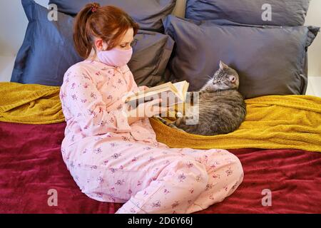 Une femme lit un livre à un chat pendant une maladie du coronavirus. Fille aux cheveux rouges en pyjama sur le lit pendant la quarantaine à la maison à côté de l'animal de compagnie Banque D'Images