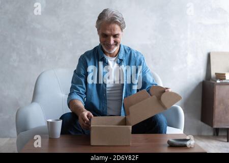 Sourire homme consommateur ouvrir boîte carton obtenir colis postal, client mâle recevoir paquet de carton s'asseoir sur le canapé à la maison Banque D'Images