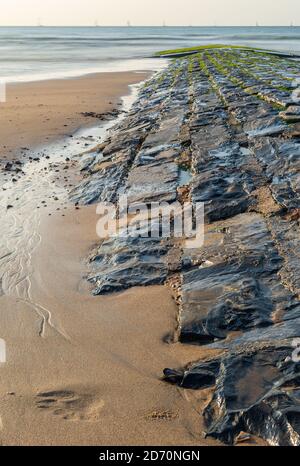 Vue verticale d'un brise-lames en pierres et en arrière-plan une mer douce avec des bateaux à voile en mouvement flou. Banque D'Images