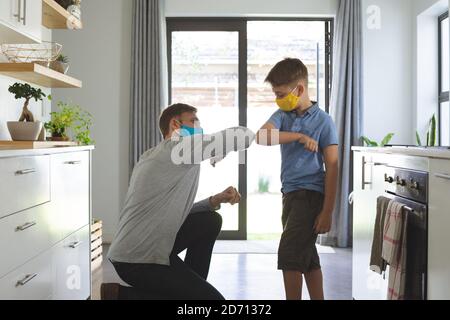 Homme caucasien à la maison avec son fils, dans la cuisine, portant des masques, saluant touchant les coudes. Distanciation sociale pendant la quarantaine du coronavirus Covid 19 Banque D'Images