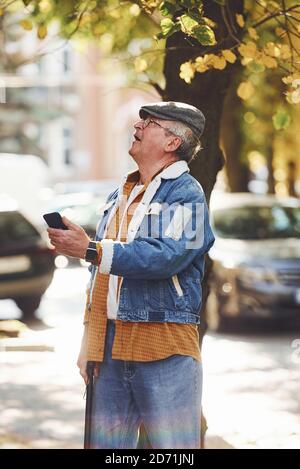 Elégant senior dans des vêtements tendance et dans des supports de lunettes avec téléphone dans la ville Banque D'Images