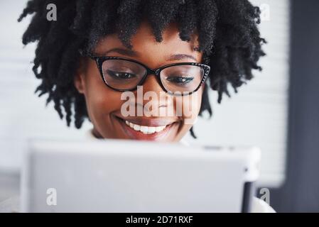 La jeune femme afro-américaine en lunettes se tient à l'intérieur dans le bureau avec tablette à portée de main Banque D'Images