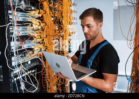 Jeune homme en uniforme et avec ordinateur portable travaille avec Internet équipement et câbles dans la salle des serveurs Banque D'Images