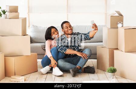 Souriant noir gars et sa petite amie se déplaçant à leur propre maison ensemble, prenant selfie parmi les boîtes de carton, panorama Banque D'Images