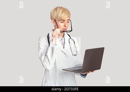 Surprise femme moyenne d'âge médecin en manteau blanc avec stéthoscope elle prend ses lunettes et regarde son ordinateur portable Banque D'Images