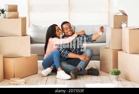 Homme noir positif et sa femme se déplaçant ensemble dans leur propre appartement, emportant le selfie parmi des boîtes avec des effets personnels Banque D'Images
