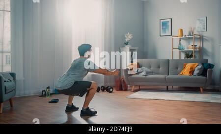 L'homme puissant Athletic Fit en T-shirt et short fait des exercices de squat à la maison dans son appartement spacieux et lumineux avec intérieur minimaliste. Banque D'Images