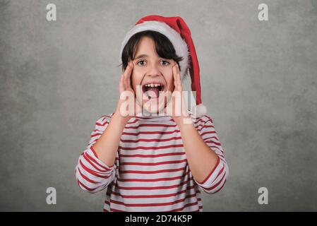 Joyeux Noël, gamin hurlant portant le chapeau du Père Noël sur fond gris Banque D'Images