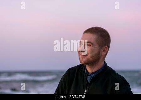 Jeune homme caucasien souriant avec une barbe sur fond de mer et des vagues avec de la mousse blanche. Mise au point sélective, gros plan. Banque D'Images