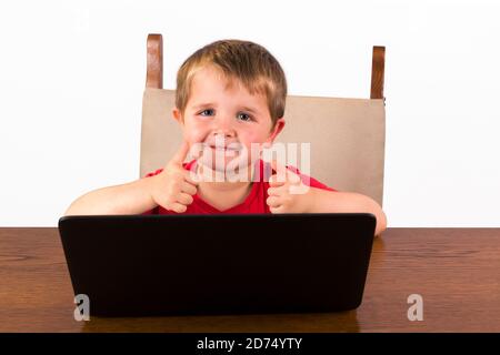 Mignon petit enfant de 4 ans assis derrière un ordinateur portable, deux pouces en regardant l'appareil photo et en souriant. Isolé sur fond blanc Banque D'Images