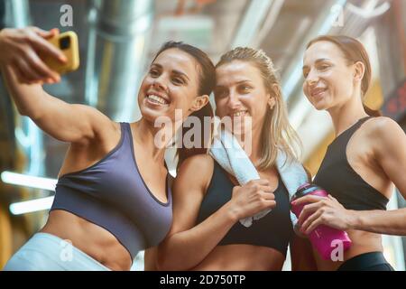 Des moments heureux. Groupe de trois jeunes femmes jeunes, belles et gaies, sportives, prenant le selfie sur un smartphone tout en se reposant après l'entraînement, s'exerçant ensemble à la salle de gym. Sport, bien-être et mode de vie sain Banque D'Images