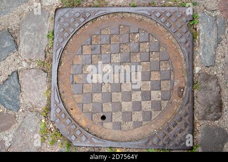 Puits d'égout de drainage de ville avec un couvercle en métal ondulé brun avec un motif en chessboard avec un coin ébréché, parmi les pierres grises de la chaussée Banque D'Images