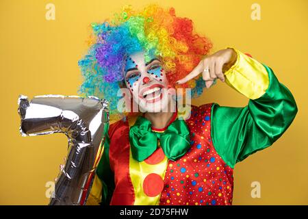 Clown se tenant sur fond jaune insolé et tenant un ballon numéro sept. Anniversaire Banque D'Images