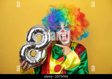 Clown se tenant sur fond jaune insolé et tenant un ballon numéro huit. Anniversaire Banque D'Images